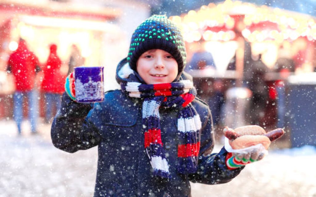Little cute kid boy eating German sausage and drinking hot children punch on Christmas market. Happy child on traditional family market in Germany. Laughing boy in colorful winter clothes.