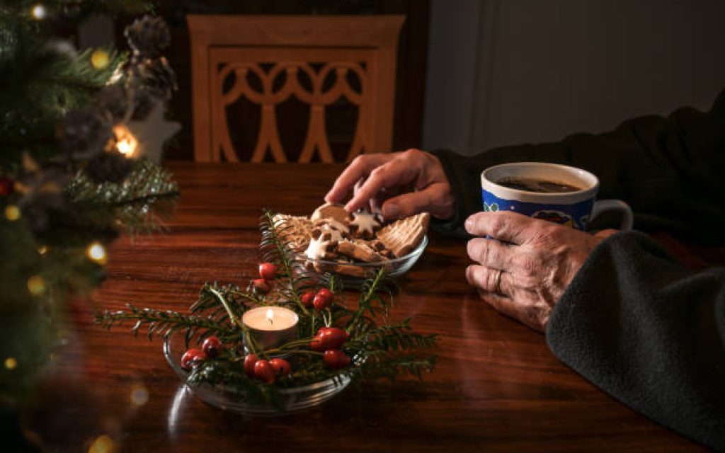 Hands of an elderly single man sitting alone at a table with Christmas cookies, coffee and festive decoration next to an empty chair, lonely holidays during the croronavirus pandemic or after a loss, selected focus
