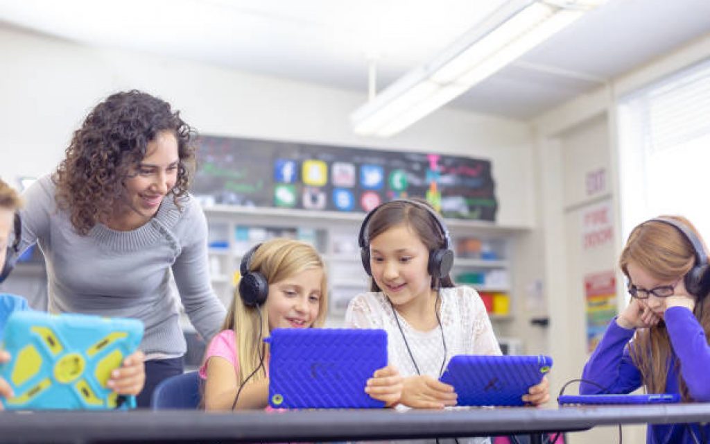 A group of elementary age students sit at their school desks and smile while using tablet computers with headphones to complete an assignment in class. A young female teacher is checking the school children's progress and providing assistance.